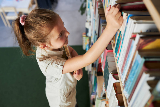School girl looking at bookshelf in school library. Smart girl selecting literature for reading. Books on shelves in bookstore. Learning from books. Back to school. Elemntary education. Benefits of everyday reading. Curious child - Фото, изображение