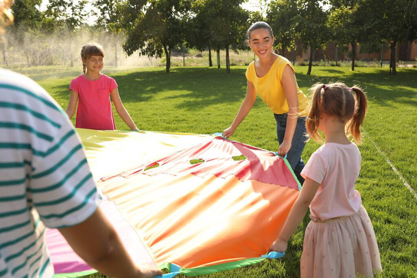Group of children and teacher playing with rainbow playground parachute on green grass. Summer camp activity - Φωτογραφία, εικόνα