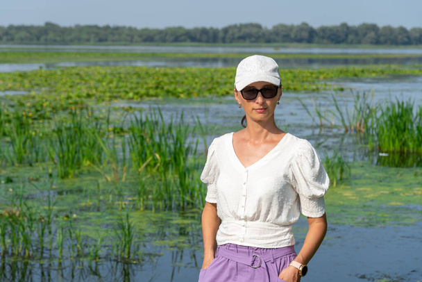 Portrait of woman on the background overgrown river. She is wearing white blouse and cap. Slowly flowing river is overgrown with yellow water lilies. Ecological problem. - Foto, Imagen