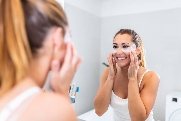 young woman after shower looking in mirror pampering herself applying face mask use anti-aging beauty product feeling happy, skin care, everyday morning routine concept - Фото, зображення