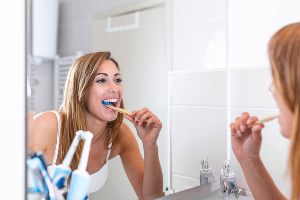 Egy vonzó nő portréja, aki fogat mos a fürdőszobában, és tükörbe néz a tükörben. egészséges fogak. Boldog szép fiatal nő néz a tükör és fogmosás a fürdőszobában - Fotó, kép
