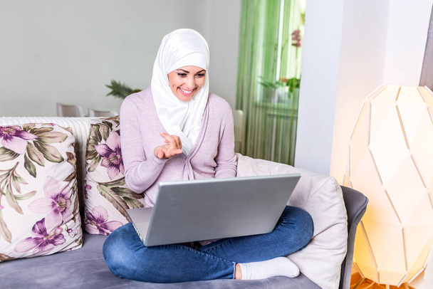 Молода мусульманка має відео-дзвінок через ноутбук вдома. Щаслива мусульманка, що сидить на дивані, сидить на дивані і користується ноутбуком у вітальні, вивчає мову, відео дзвінок. - Фото, зображення