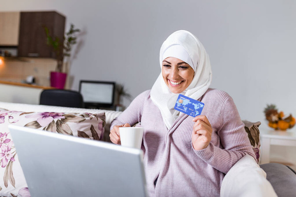 Κομψό ελκυστική μουσουλμάνα γυναίκα που χρησιμοποιεί κινητό φορητό υπολογιστή ψάχνει σε απευθείας σύνδεση πληροφορίες για ψώνια στο σαλόνι στο σπίτι. Πορτρέτο της ευτυχούς γυναίκας που αγοράζει το προϊόν μέσω online αγορών. πληρώνουν με πιστωτική κάρτα - Φωτογραφία, εικόνα
