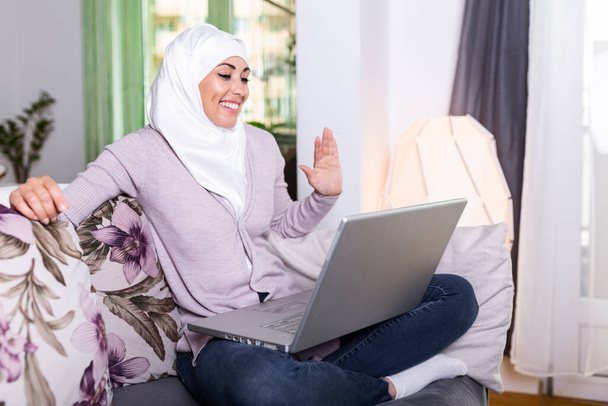 Junge Muslimin telefoniert zu Hause per Laptop. Glücklich lächelnde Muslimin auf Sofa, Couch und Laptop zu Hause im Wohnzimmer, Sprache lernen, Videotelefonie. - Foto, Bild