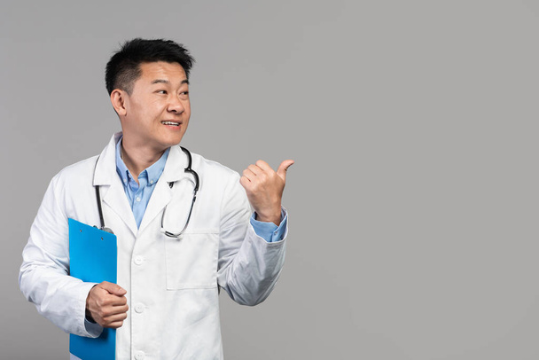 Lächelnder selbstbewusster erwachsener asiatischer Arzt in weißem Kittel mit Stethoskop und Tablette, der mit dem Finger auf den leeren Raum zeigt, isoliert auf grauem Hintergrund, Studio. Medizinischer Service, Gesundheitsfürsorge, tolles Angebot und Werbung - Foto, Bild