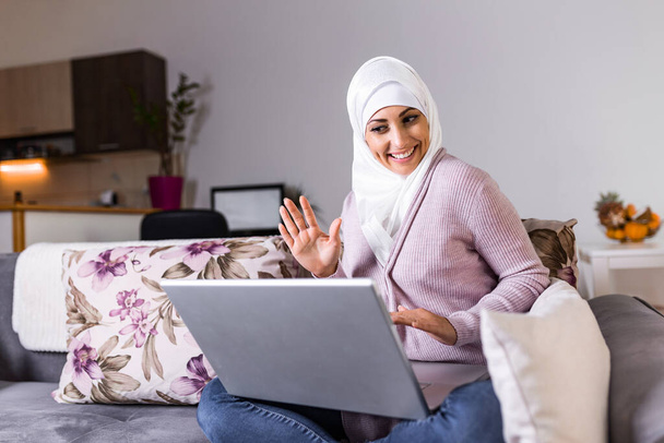 Μουσουλμάνα που δουλεύει στον υπολογιστή. Αραβα Νεαρή επιχειρηματίας κάθεται στο γραφείο της στο σπίτι, εργάζεται σε ένα φορητό υπολογιστή και πίνοντας καφέ ή τσάι. Μουσουλμάνα γυναίκα που εργάζεται σε σπίτι και χρησιμοποιεί υπολογιστή. - Φωτογραφία, εικόνα