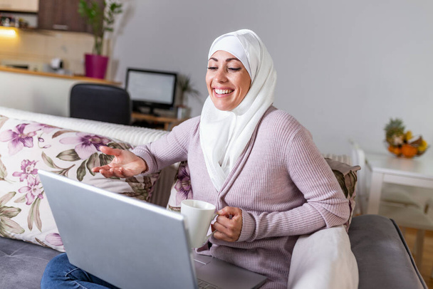 Young Muslim woman having video call via laptop at home. Happy smiling Muslim woman sitting on sofa, couch and using laptop at living room at home, learning language, video calling. - Zdjęcie, obraz