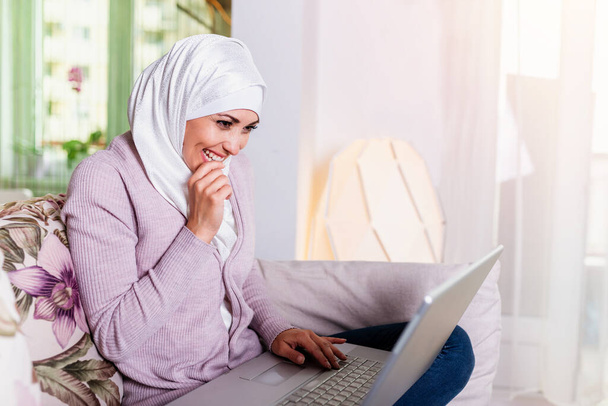 Muslim woman Using her Laptop, Surfing Social Networks at Home, Free Space. Surfing the net at home. Side view of attractive young Muslim woman working on laptop and smiling while sitting on the couch at home - Φωτογραφία, εικόνα