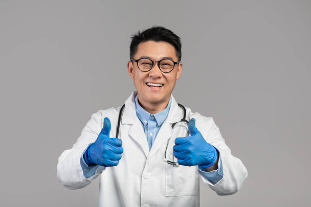 Glad middle aged japanese man doctor in white coat, glasses and protective gloves shows thumbs up, isolated on gray background, studio. Health care, therapist advice and approving modern medicine - Photo, Image