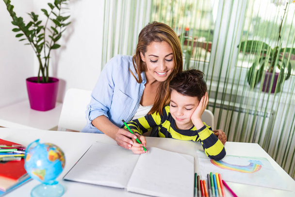 Mom helping little boy to do homework. Homeschooling during the coronavirus pandemic. Cute boy doing his school homework with his mother, at home, he is writing on a book - Фото, изображение