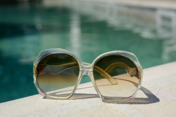 Stylish sunglasses near outdoor swimming pool on sunny day, closeup. Beach accessory - Fotó, kép