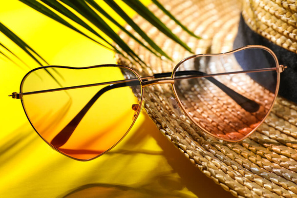 Stylish heart shaped sunglasses and straw hat on yellow background, closeup - Zdjęcie, obraz