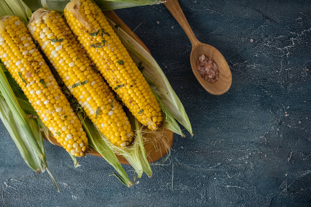 Grilled corn on the cob served in the wooden plate with salt. Dark concrete background. Yellow corn head. Flat lay - Foto, Bild
