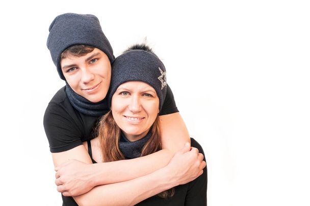 A teenager wearing a warm hat and scarf on his neck embraces his mother, who is also dressed in a hat on a white background. Son and mom are glad that they will not be cold in winter. Isolated - Foto, Imagem