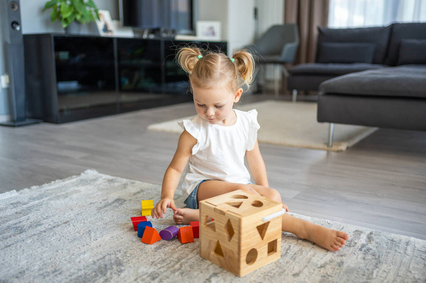 Nettes kaukasisches kleines Mädchen, das zu Hause mit Öko-Holzspielzeug auf dem Boden spielt. Montessori-Spielzeug. Das Kind, das Lernspiele spielt. Hochwertiges Foto - Foto, Bild