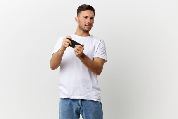 Homem bonito bronzeado feliz na t-shirt básica jogar jogo online com telefone posando isolado sobre fundo estúdio branco. Copiar espaço Banner Mockup. Conceito de RPG de jogos móveis - Foto, Imagem