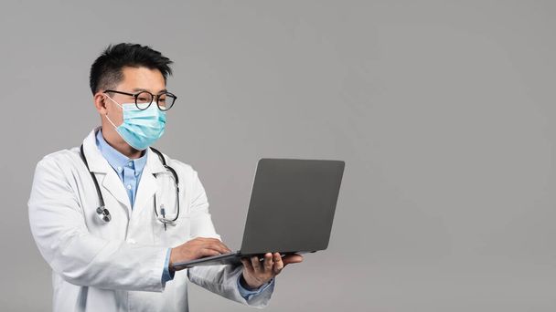 Confident adult chinese man doctor in white coat, glasses, protective mask typing on laptop isolated on gray background. Medicine, health care, social distance and online consultation due covid-19 - Фото, изображение