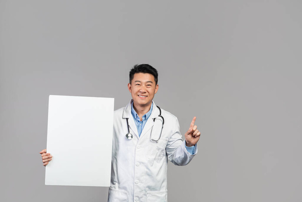 Glücklich reifer japanischer Arzt in weißem Mantel mit Stethoskop und Banner mit Leerraum, der den Finger nach oben zeigt, isoliert auf grauem Hintergrund, Studio. Medizinischer Dienst, Gesundheitsfürsorge, Beratung, Anzeige und Angebot - Foto, Bild