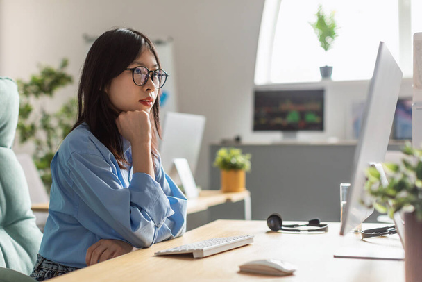 Junge koreanische Geschäftsfrau, die online am Computer im Internet arbeitet, sitzt im modernen Büro und trägt eine Brille. Unternehmenskarriere und weibliches Unternehmertum. Seitenansicht - Foto, Bild