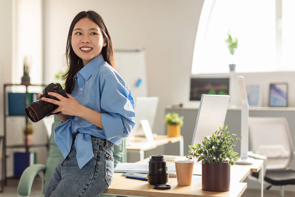 Cheerful Japanese Millennial Photographer Woman Holding Photocamera Posing Looking Aside In Modern Office. Creative Digital Photography Art And Career Concept - 写真・画像