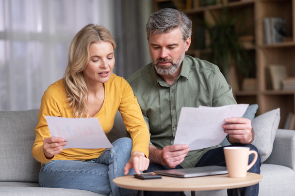Financial Literacy. Busy Middle Aged Husband And Wife Calculating Family Budget Together While Sitting On Couch In Living Room Interior, Spouses Checking Loan Documents And Using Calculator - 写真・画像