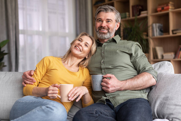 Smiling Married Middle Aged Couple Spending Time At Home Together, Drinking Coffee And Watching Tv, Cheerful Spouses Relaxing On Couch In Living Room, Enjoying Domestic Pastime, Closeup Shot - Foto, imagen