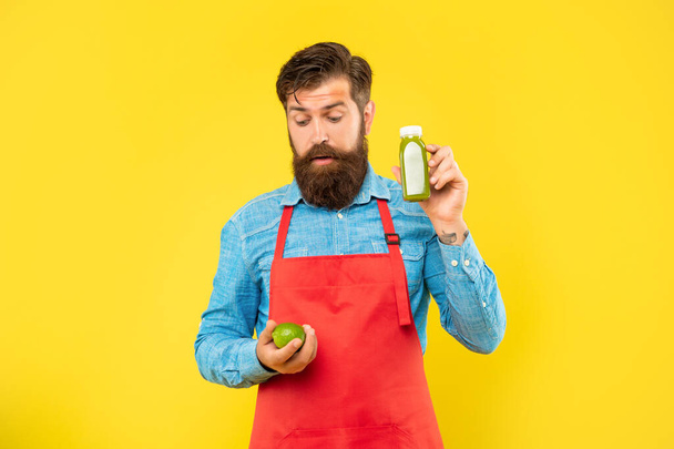 Surprised man in apron looking at limes holding juice bottle yellow background, juice shopkeeper. - Photo, image