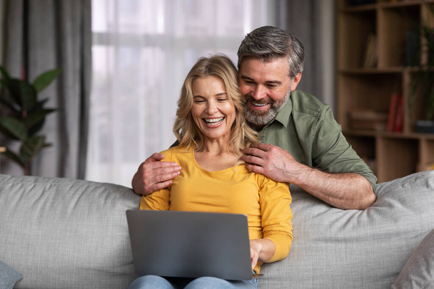 Happy Middle Aged Couple With Laptop Having Fun Together At Home, Cheerful Spouses Using Computer While Relaxing On Couch In Living Room, Browsing Internet Or Shopping Online, Copy Space - 写真・画像