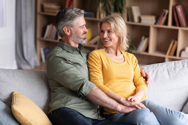 Happy Marriage. Portrait Of Smiling Middle Aged Spouses Embracing On Couch At Home, Cheerful Married Couple Resting On Sofa In Living Room, Loving Man And Woman Hugging And Looking At Each Other - Foto, Imagen
