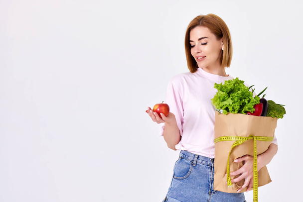 young caucasian woman in pink t-shirt hold paper bag with groceries and with measuring tape and red apple on white background. copy space - Fotoğraf, Görsel
