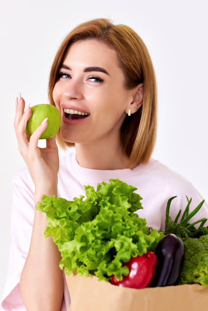 smiling woman holding paper bag with vegetables and biting green apple on white background. close-up - Fotoğraf, Görsel