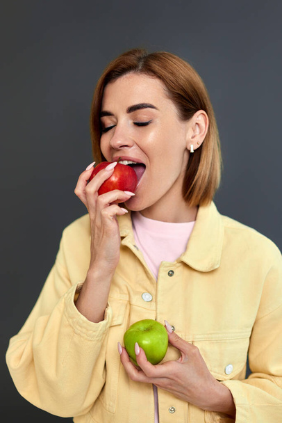 smiling blonde woman holding green and red apples on yellow background. - Fotografie, Obrázek