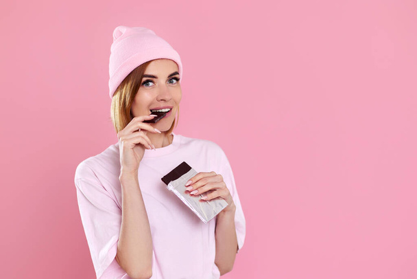 young happy woman in casual t-shirt biting delicious chocolate bar and looking at camera isolated on pastel pink background - Valokuva, kuva