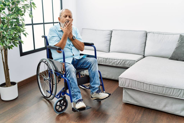 Handsome senior man sitting on wheelchair at the living room bored yawning tired covering mouth with hand. restless and sleepiness.  - Foto, immagini