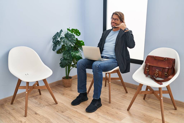 Handsome middle age man sitting at waiting room working with laptop smiling doing phone gesture with hand and fingers like talking on the telephone. communicating concepts.  - 写真・画像