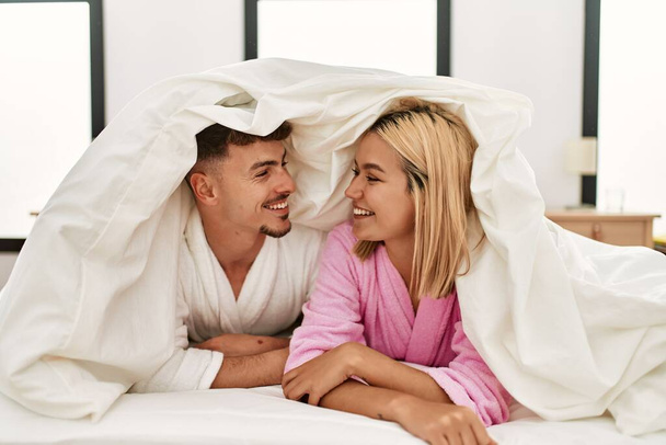 Jeune couple caucasien couvrant avec drap de lit et souriant heureux couché sur le lit à la maison. - Photo, image