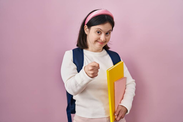 Woman with down syndrome wearing student backpack and holding books pointing fingers to camera with happy and funny face. good energy and vibes.  - Φωτογραφία, εικόνα
