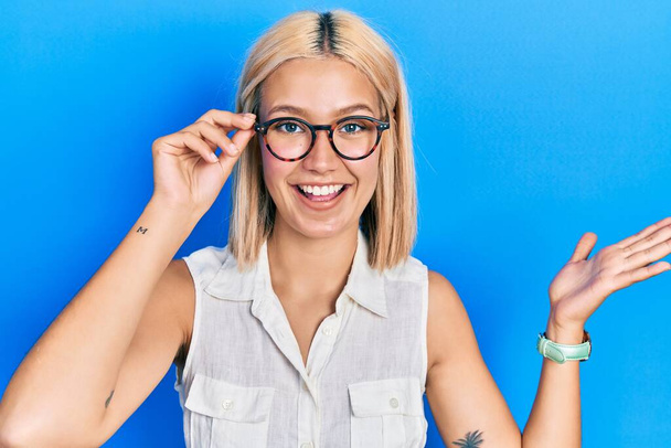 Beautiful blonde woman wearing glasses celebrating achievement with happy smile and winner expression with raised hand  - Valokuva, kuva