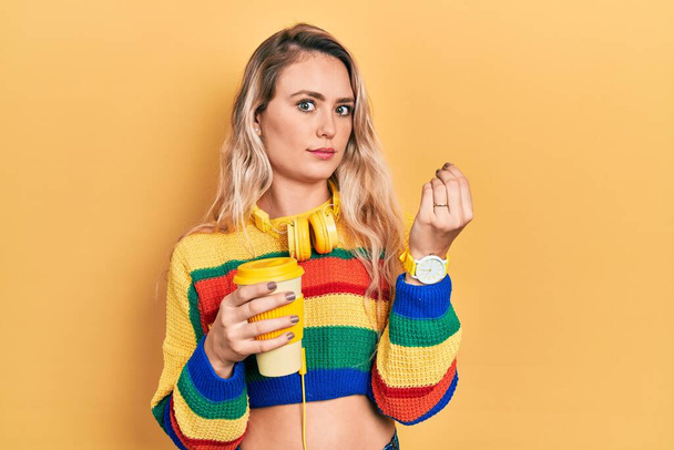 Beautiful young blonde woman drinking cup of coffee wearing headphones doing italian gesture with hand and fingers confident expression  - Zdjęcie, obraz