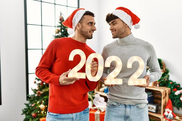 Two hispanic men couple celebrate new year 2022 standing by christmas tree at home - Photo, image
