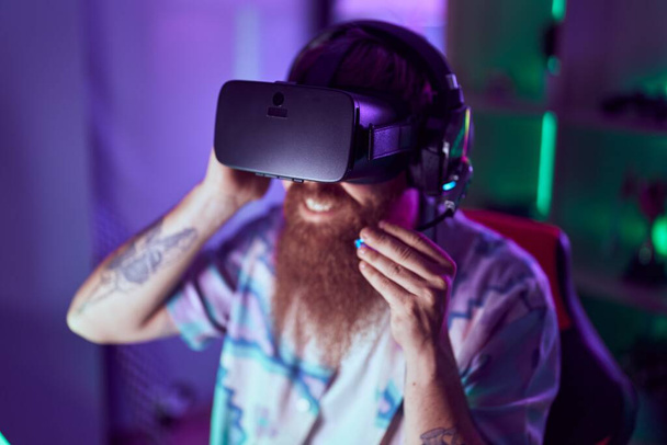 Young redhead man streamer playing video game using virtual reality glasses at gaming room - Foto, Bild