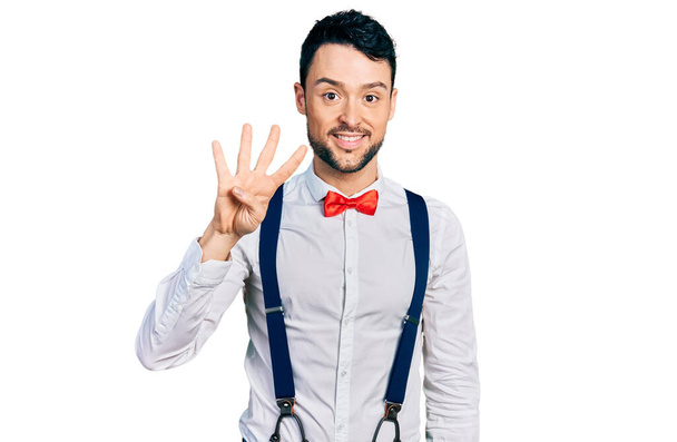 Homme hispanique avec barbe look hipster avec noeud papillon et bretelles montrant et pointant vers le haut avec les doigts numéro quatre tout en souriant confiant et heureux.  - Photo, image