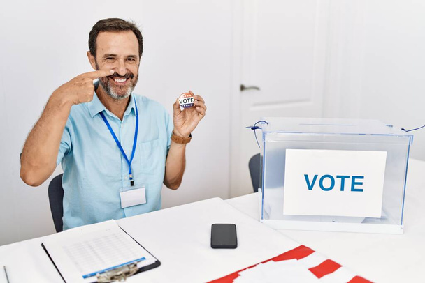 Чоловік середнього віку з бородою, сидячи за голосуванням, тримає я голосую значок, вказуючи ручним пальцем на обличчя і ніс, усміхаючись весело. концепція краси
  - Фото, зображення