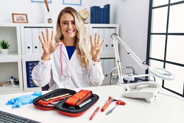 Young beautiful doctor woman with reflex hammer and medical instruments showing and pointing up with fingers number nine while smiling confident and happy.  - 写真・画像