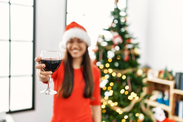 Fiatal latin nő iszik bort álló karácsonyfa otthon - Fotó, kép