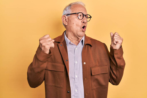 Senior man with grey hair wearing casual jacket and glasses showing arms muscles smiling proud. fitness concept.  - Foto, imagen
