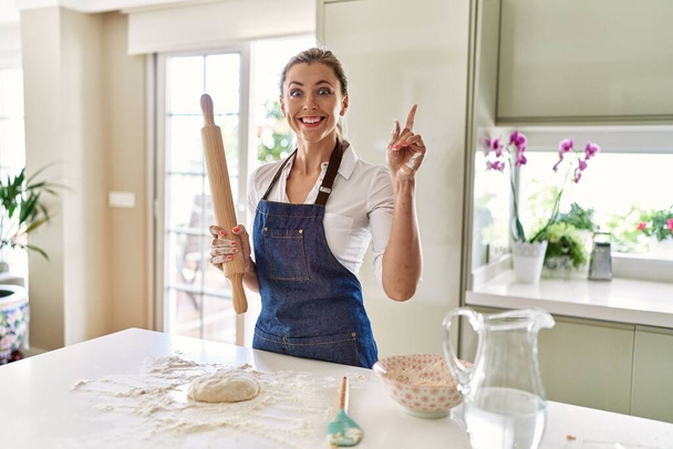 Beautiful blonde woman preparing dough for pizza surprised with an idea or question pointing finger with happy face, number one  - Photo, image