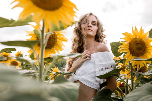 a young girl with a model appearance, blond and long hair, in denim trousers and a white blouse in a sunflower field. High quality photo - Photo, Image