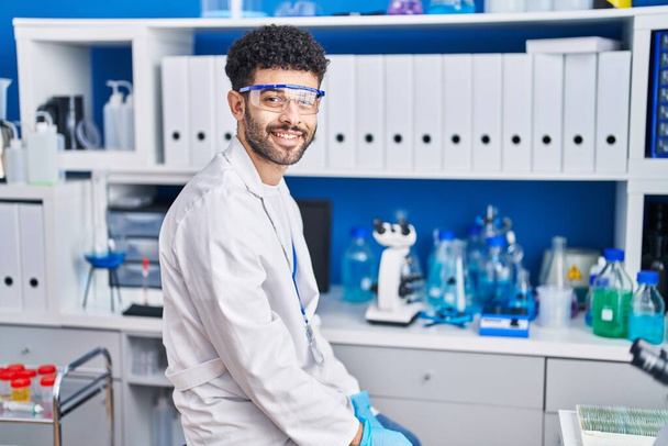 Fiatal arab férfi, tudós egyenruhát visel, magabiztosan mosolyog a laboratóriumban. - Fotó, kép