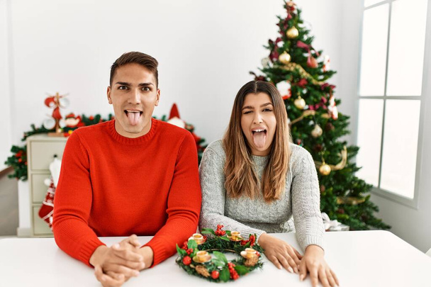 Молодая испаноязычная пара, сидящая за столом на Рождество, торчащая языком, довольная смешным выражением лица. концепция эмоций.  - Фото, изображение